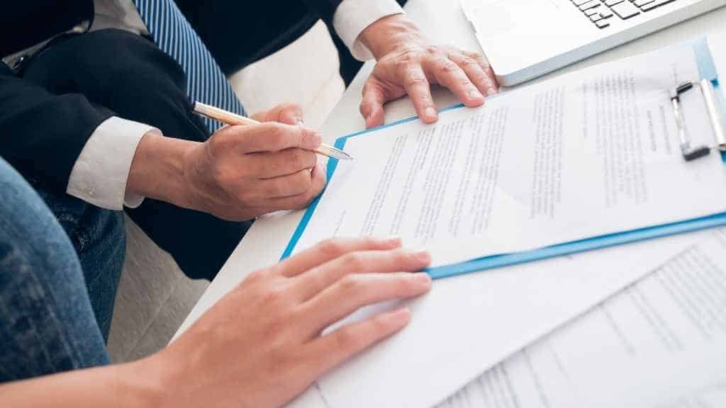 Ein Vertrag, der von einer Person unterschrieben wird. Die Hand einer zweiten Person hält den Vertrag.