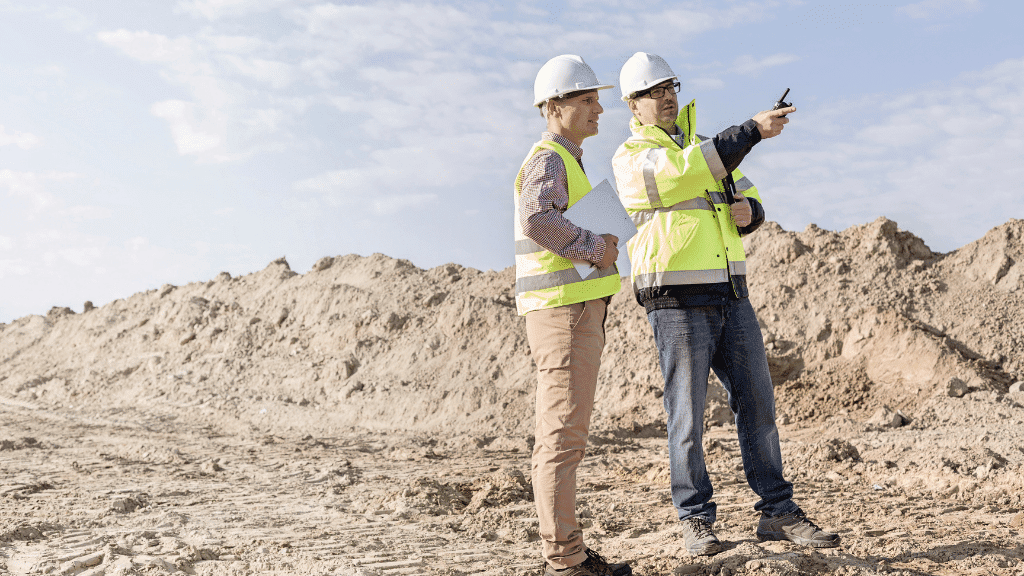 Zwei Bauarbeiter mit Helm und Warnwesten stehen auf einer Baustelle.