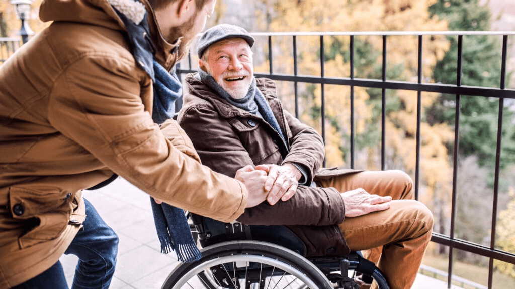 Ein älterer Mann sitzt im Rollstuhl und ein junger Mann gibt ihm die Hand.